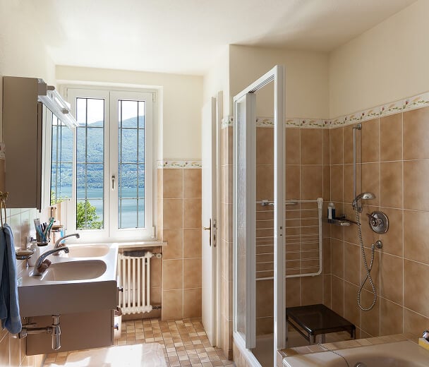 Bright bathroom with beige tiles, a shower, and a white sink. Sunlight streams through a window, revealing a scenic view of mountains and water.