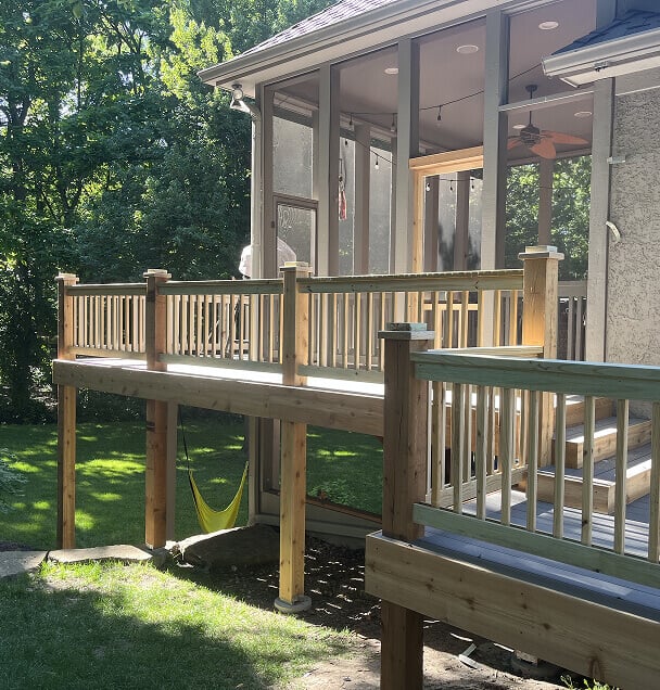 A sunlit wooden deck extends from a house, featuring a fenced railing and steps. A yellow hammock hangs beneath. Lush green trees surround the area.