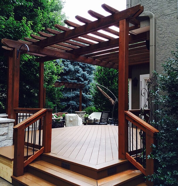 A wooden deck with a pergola leads to a garden area. Stairs and railings frame the deck, surrounded by lush greenery, creating a cozy, inviting ambiance.