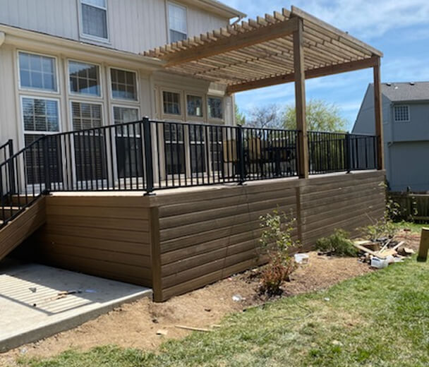 A spacious wooden deck with black metal railings and a pergola, attached to a house with large windows. The setting is bright and sunny.