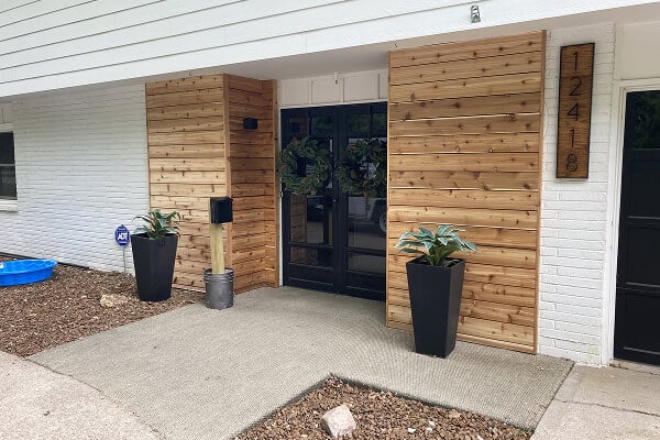 Modern house entrance with black double doors flanked by wood paneling and tall planters. House number "12418" is displayed on the right.