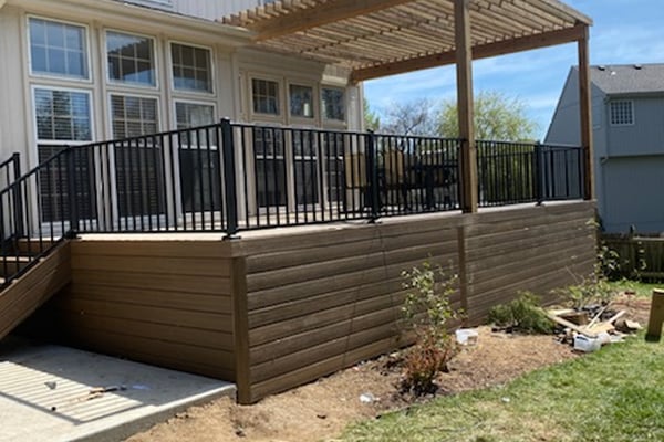 Wooden deck with black railings and a pergola, attached to a house with large windows. The area beneath is landscaped with young plants. Bright, sunny day.