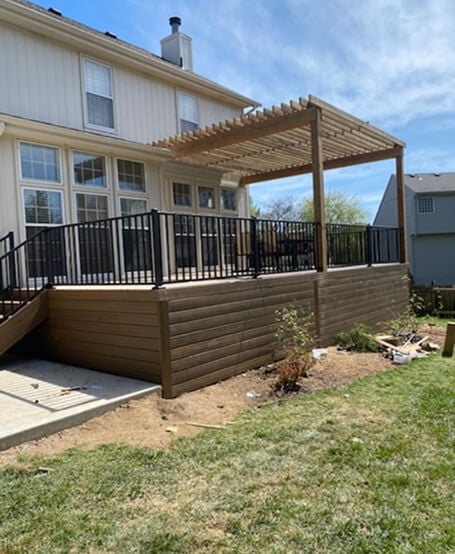 Two-story house with a wooden deck and black railings. A pergola provides shade. The yard is partly landscaped, reflecting a calm, suburban setting.