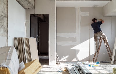 A construction worker on a ladder applies plaster to drywall in a partially finished room. Tools and materials are scattered on the floor, suggesting ongoing renovation.