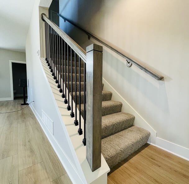 A carpeted staircase with wooden handrails and black metal spindles ascends in a modern interior. Light wood flooring and neutral walls enhance the space.