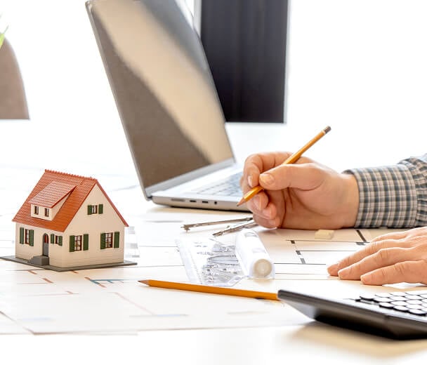 A person is working on architectural plans at a desk, with a laptop, calculator, and a small model of a house nearby.