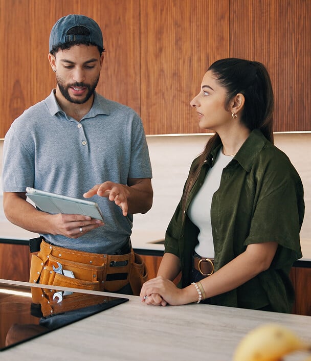 A man in a grey polo shirt and cap is showing something on a tablet to a woman in a green oversized shirt, both are engaged in conversation in a modern kitchen setting.