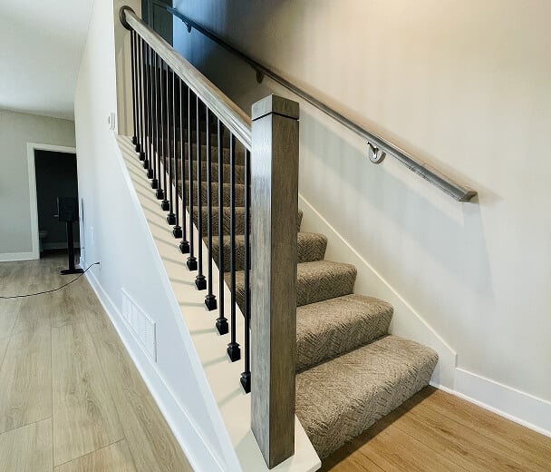 A carpeted staircase with wooden handrails and black metal spindles ascends in a modern interior. Light wood flooring and neutral walls enhance the space.