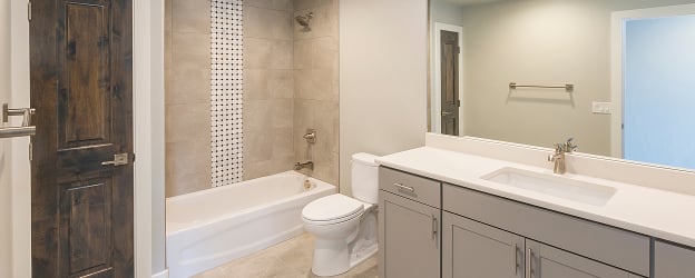 A modern bathroom featuring a bathtub with a tile accent wall, a toilet, and a vanity with a sink and cabinetry. There is also a mirror above the vanity and neutral-colored walls.