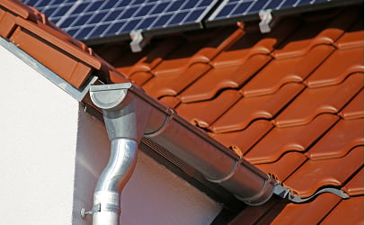 Close-up of a red-tiled roof with solar panels partially visible. A shiny metal gutter system runs along the edge, evoking a modern, eco-friendly home.