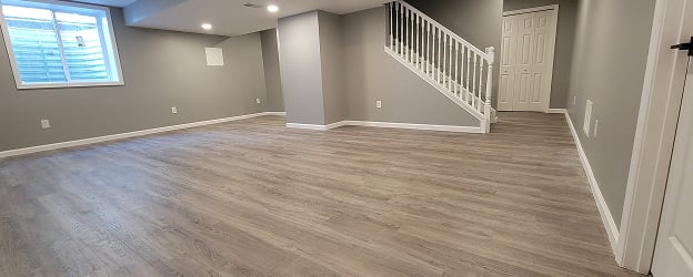 Spacious basement with light gray walls, rustic wood flooring, and white trim. A staircase leads up, and a window provides natural light, creating an inviting tone.