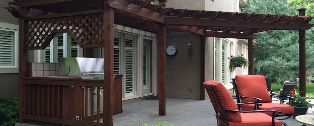 A wooden pergola covers a patio with red cushioned chairs and a built-in grill. The setting is peaceful, with greenery and a wall clock visible.