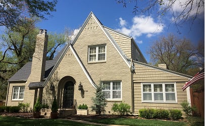 Charming beige brick house with a steep gabled roof, surrounded by green trees and shrubs. An American flag is visible on the right, under a bright blue sky.