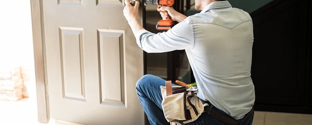 A man kneeling, using an orange drill to install a beige door. He wears a light blue shirt and jeans, with a tool belt. The scene conveys focus and craftsmanship.
