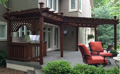 A wooden pergola covers a backyard patio with red cushion chairs and a barbecue grill. The setting is serene with lush greenery surrounding it.