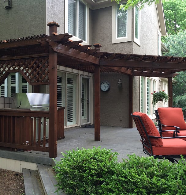 Outdoor patio with wooden pergola attached to a house. Features a grill, clock, red-cushioned chairs, and lush greenery, creating a cozy ambiance.