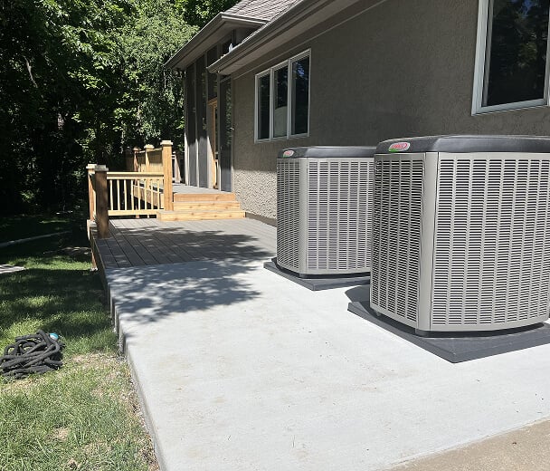 Two large air conditioning units sit on a concrete patio beside a house with beige siding. In the background is a wooden deck under trees.