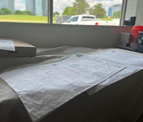 Blueprints laid out on a table by a window, with a backdrop of a park and buildings. A truck is partially visible outside. The scene is calm and studious.