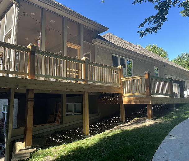 Wooden deck attached to a beige house with large windows, under a clear blue sky. Shadows from nearby trees stretch over the grass below.