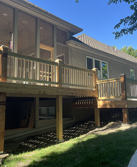 Wooden deck attached to a beige house with large windows, under a clear blue sky. Shadows from nearby trees stretch over the grass below.