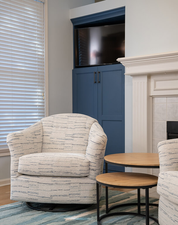 Cozy living room with a patterned armchair, round wooden nesting tables, blue cabinet with TV, and a white fireplace, creating a calm ambiance.