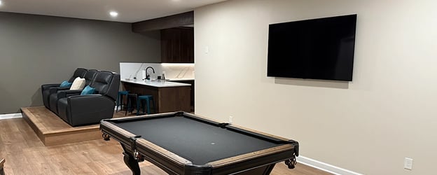 Modern entertainment room with a black pool table centered. Black recliners and a kitchenette with barstools are in the background. Wall-mounted TV on beige wall.