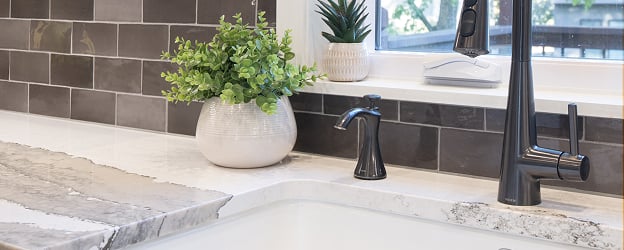 Modern kitchen sink area with sleek black faucet, white countertop, and gray tile backsplash. Decor includes potted plants for a fresh look.