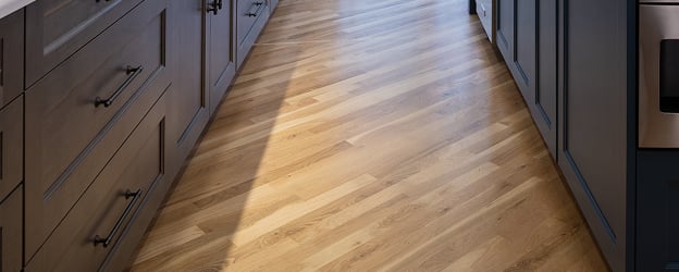 Warm wooden flooring stretches down a modern, narrow kitchen with sleek dark cabinets on both sides, creating a cozy and elegant atmosphere.