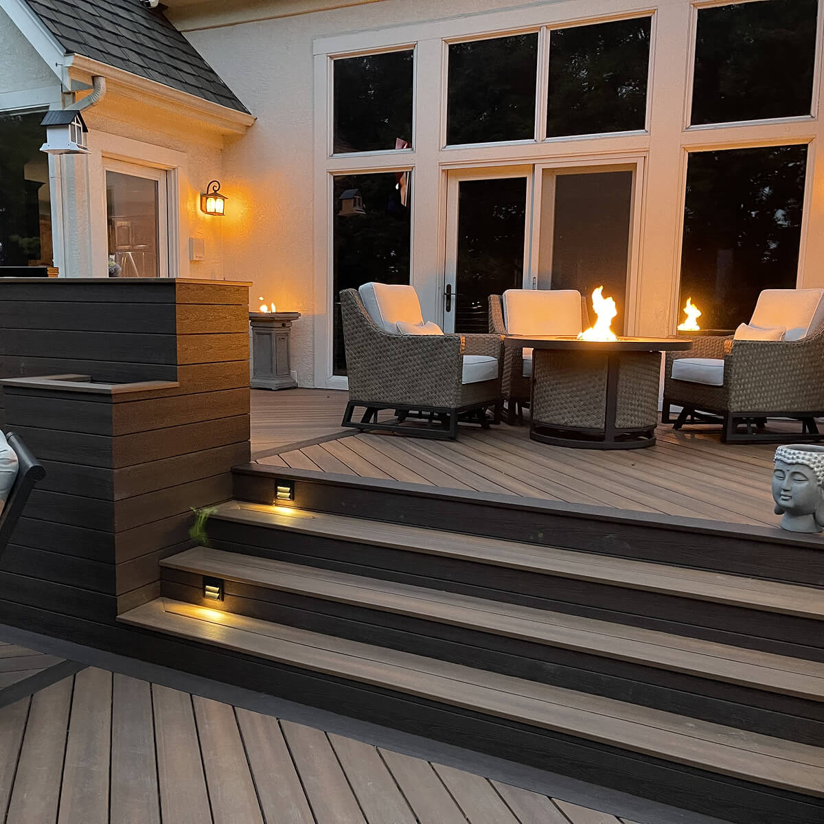 Outdoor patio with wicker chairs around a fire pit table, glowing warmly. Steps with soft lighting lead down. The setting is cozy and inviting.