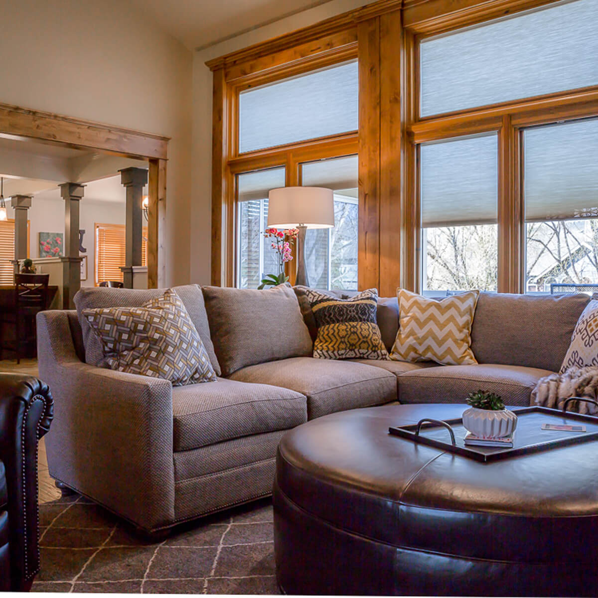 A cozy living room with a large beige sectional sofa adorned with patterned cushions. Wooden windows let in natural light, while a round leather ottoman adds elegance.