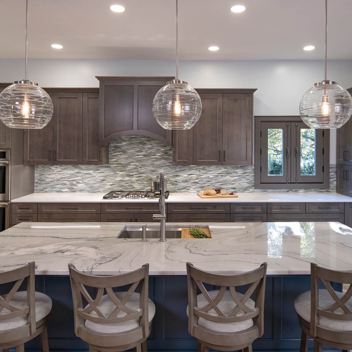 A modern kitchen featuring wooden cabinets, a large marble countertop island with bar stools, pendant lighting, and stainless steel appliances.